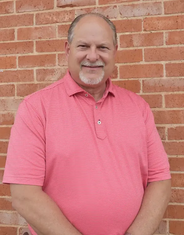 Smiling man in a pink striped polo shirt standing against a brick wall.