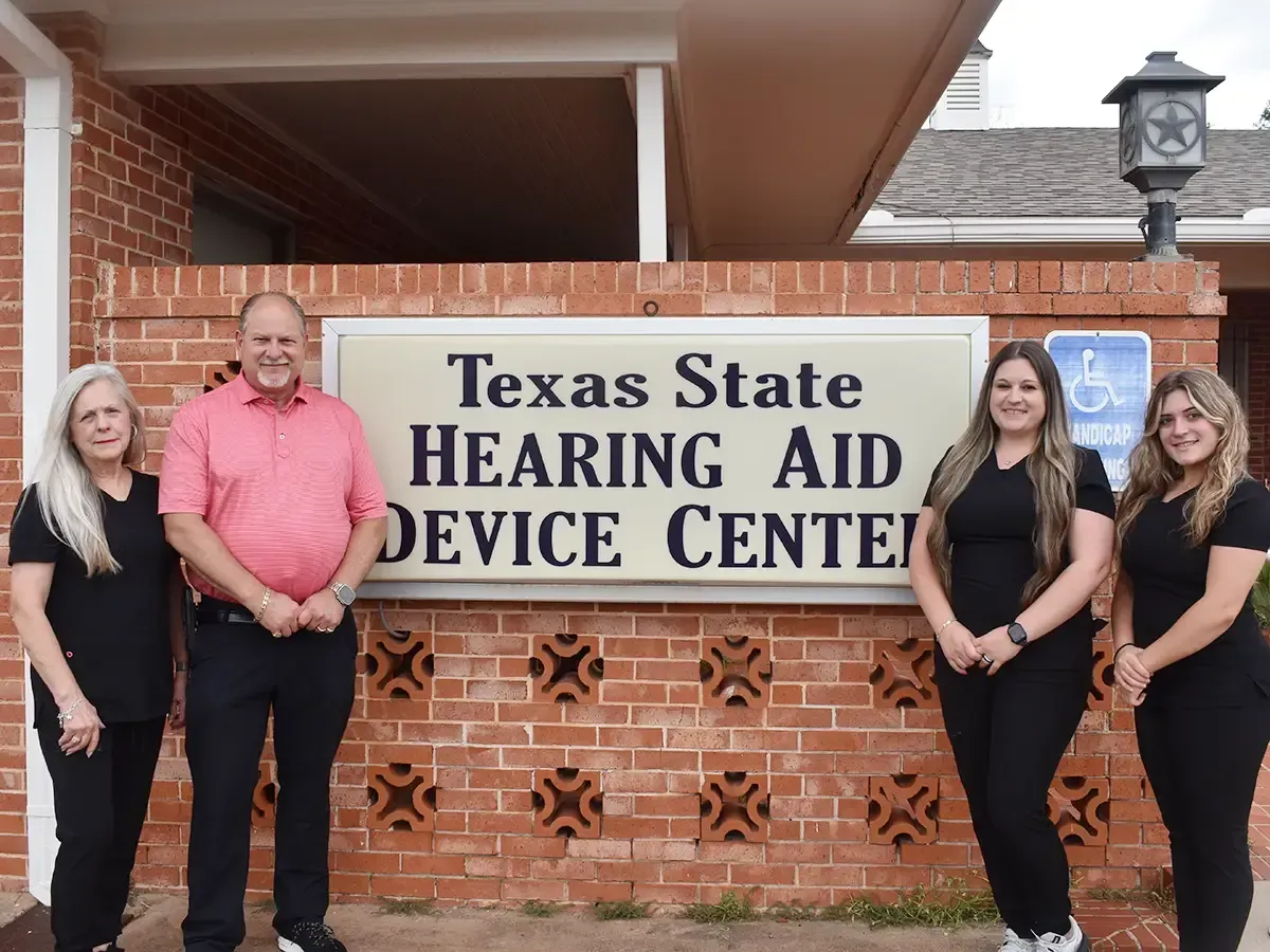 Texas State Hearing Aid Device Center sign with four people standing in front of a brick building