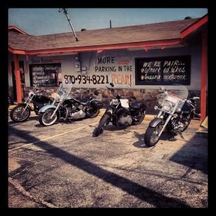 Motorcycles lined up outside a red roadside repair shop with hand-painted signs.