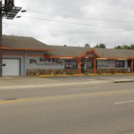 Street view of a low gray storefront with orange trim and a sign, beside a road under cloudy skies