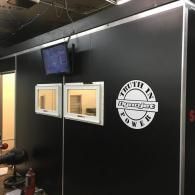 Black wall with two small windows, a monitor above, and a round white logo in an indoor workspace