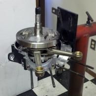 Metal machine component with circular top plate, levers, and a red stand in a workshop