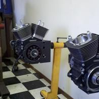 Two black V-twin engine models on display, one mounted on a yellow stand in a checkered-floor workshop.