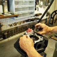 Hands working on an engine block in a workshop, using a wrench near the top cylinder opening.