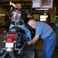 Two men working on a motorcycle inside a repair shop, one seated on the bike and one crouching beside it.