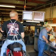Two men in a garage workshop, one on a motorcycle, both facing two computer monitors.