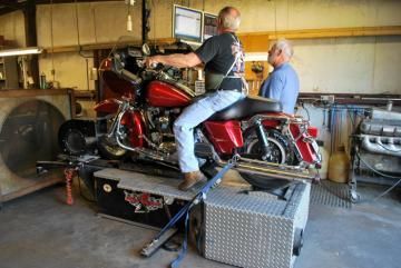 Two men on a red touring motorcycle strapped on a dynamometer in a workshop