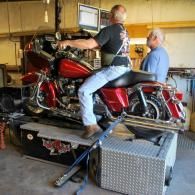 Two men load a red motorcycle onto a trailer inside a garage.