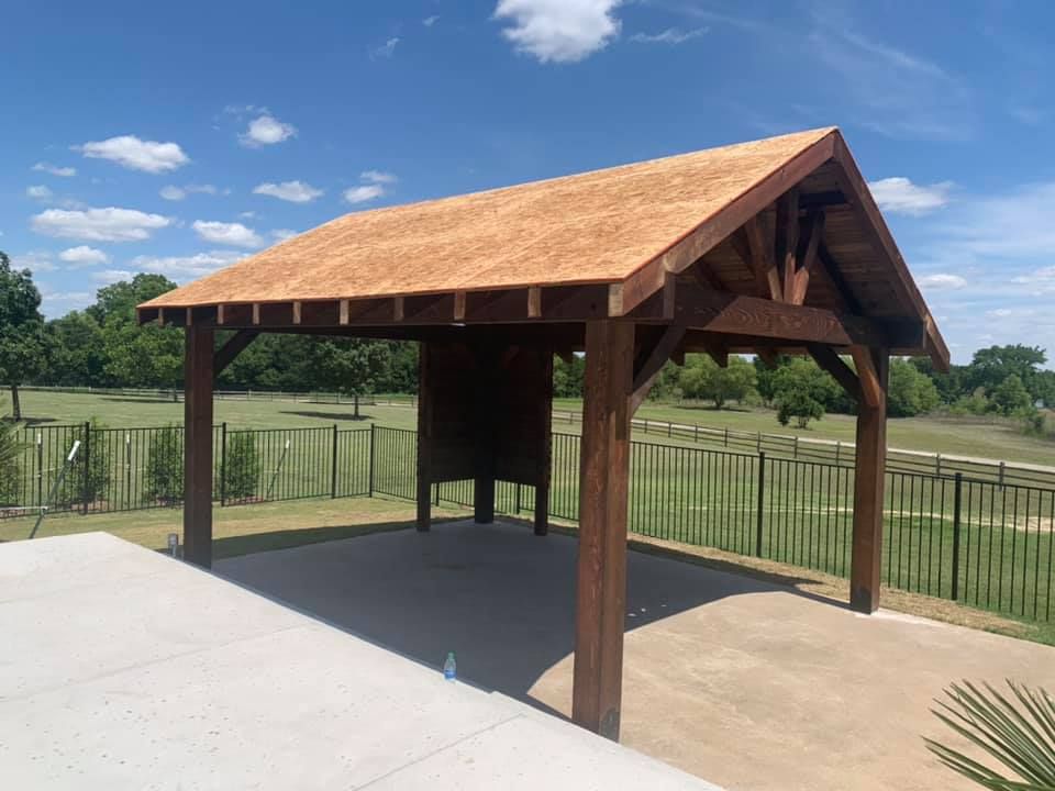 Wooden pavilion with a peaked roof on a concrete pad in a fenced park under a blue sky.