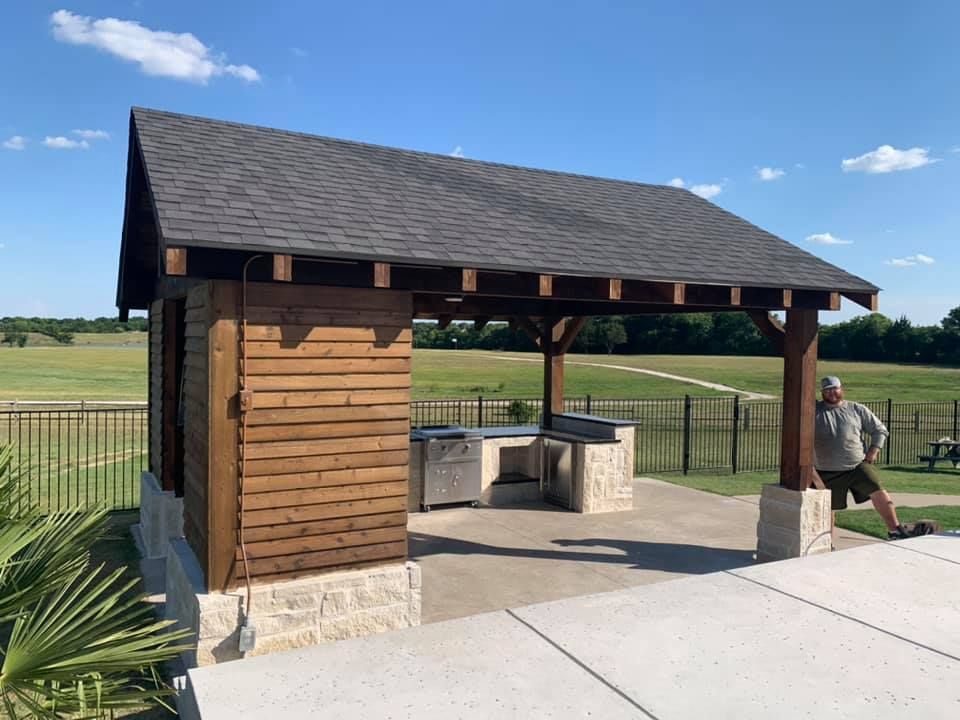 Open-sided wooden pavilion with stone counter in a grassy park, a person stands nearby