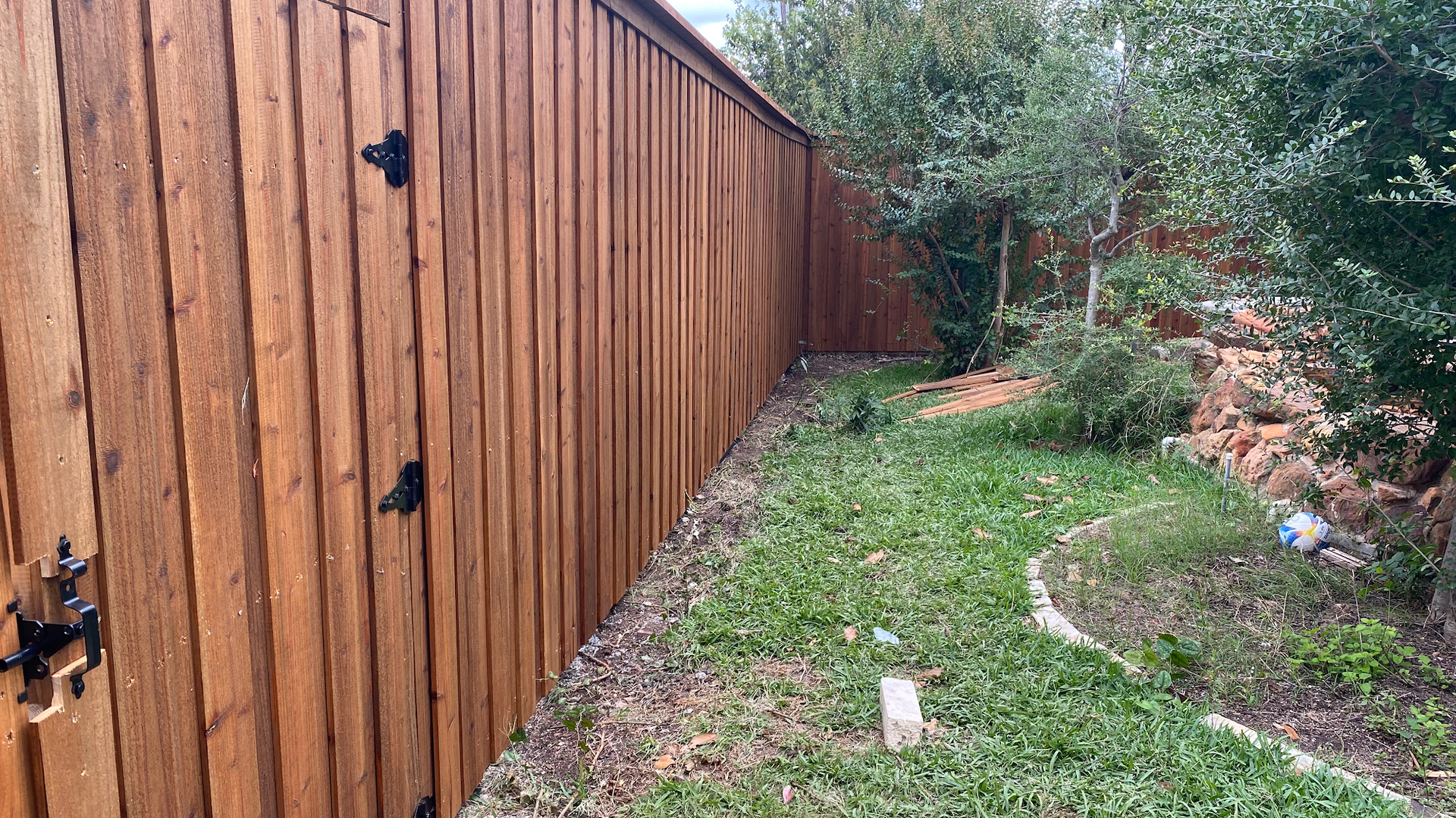 Wooden fence along a narrow grassy side yard with trees and a dirt path.