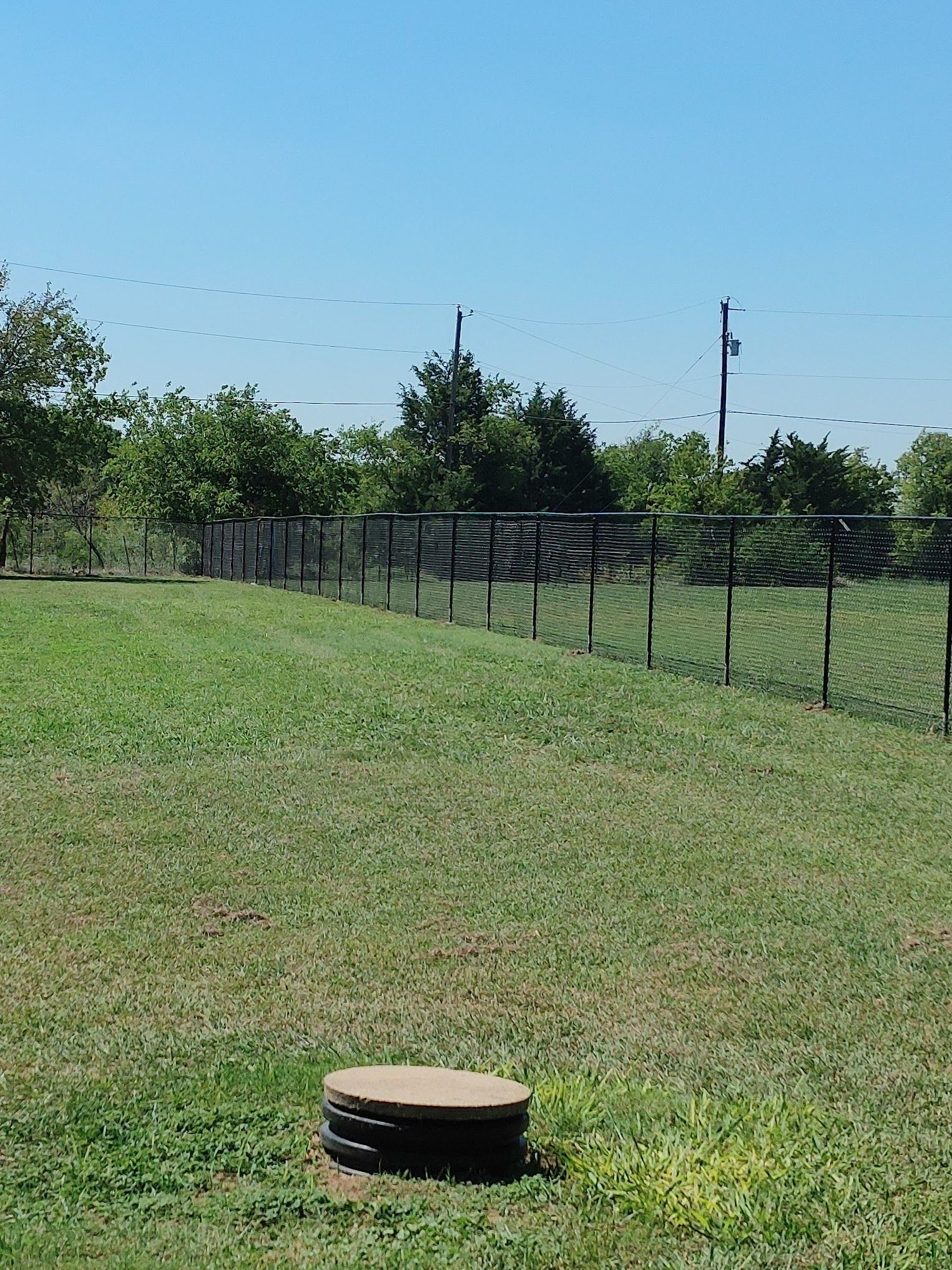 Grassy field with a chain-link fence, trees, and a round utility cover in the foreground under a blue sky
