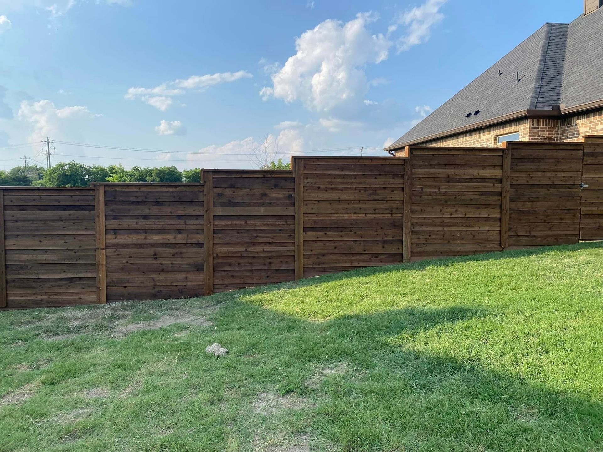 Wooden fence along a grassy backyard under a blue sky with clouds, beside a house roof.