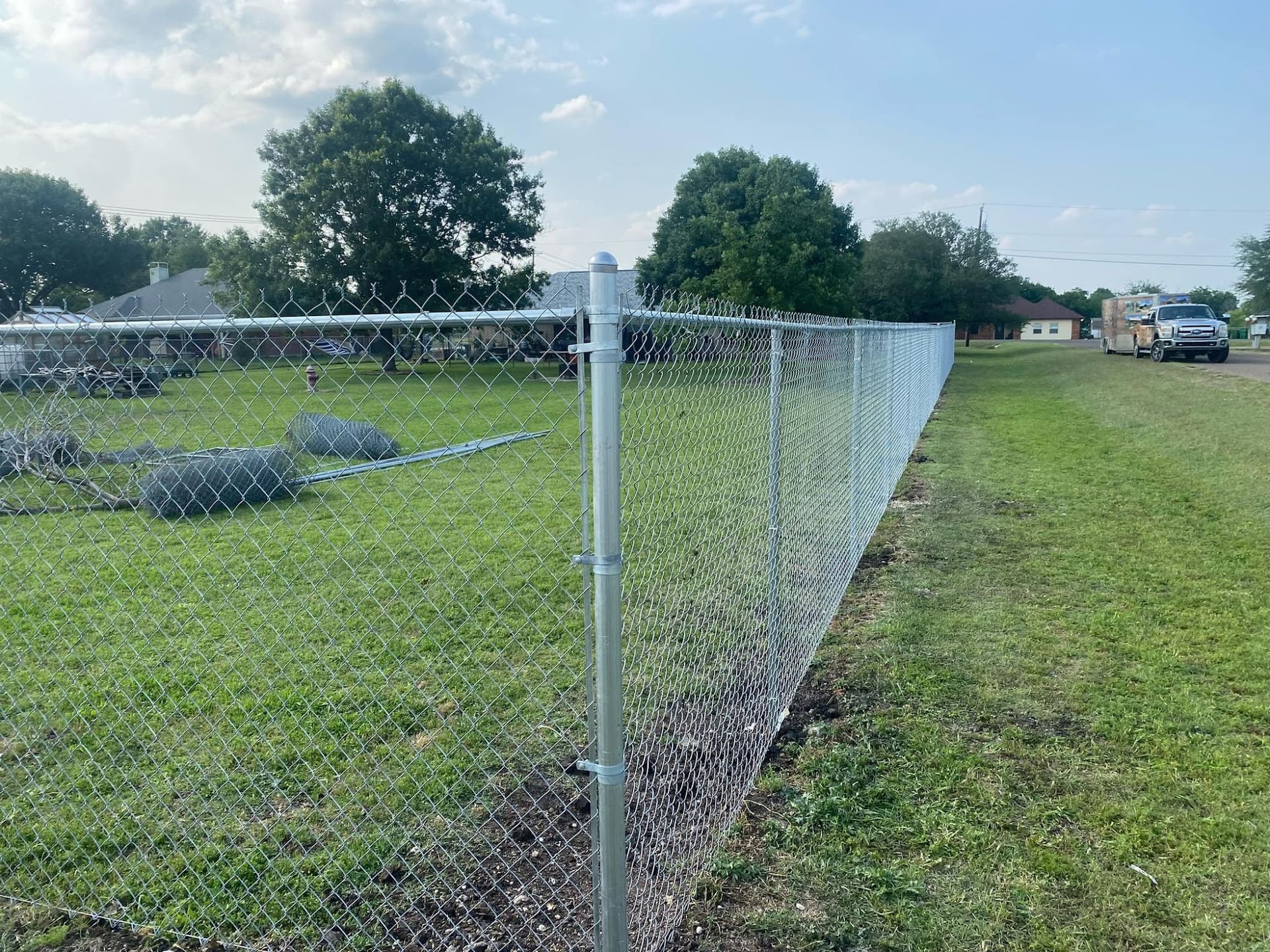 Chain-link fence along a grassy field with rocks, trees, and a cloudy sky