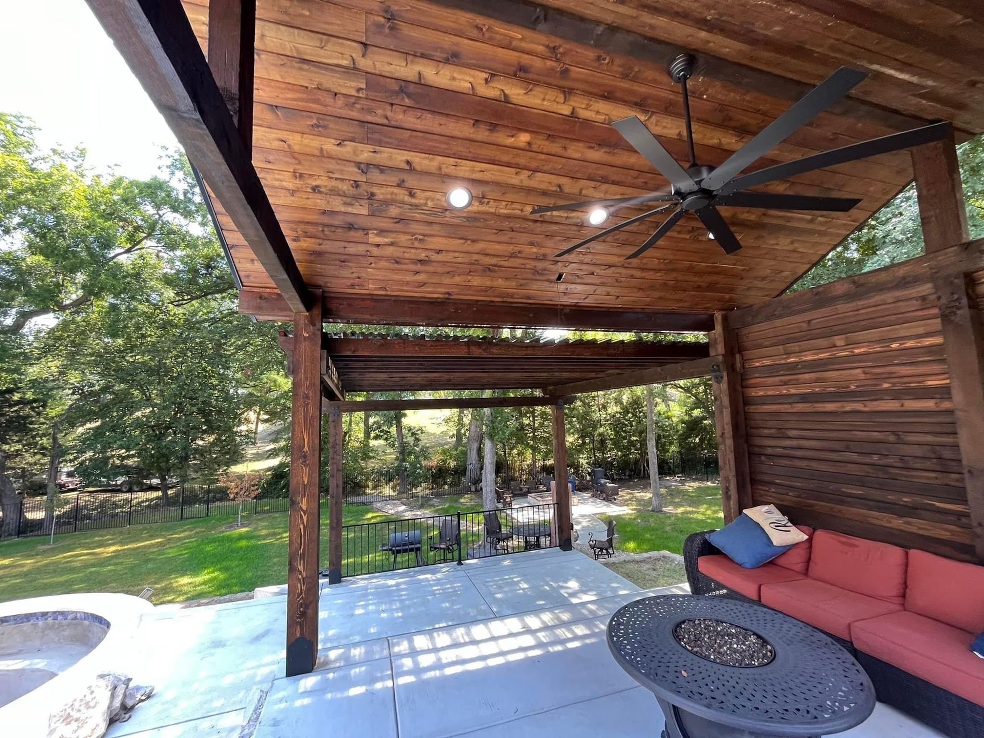 Covered wooden patio with ceiling fan, red sofa, and outdoor seating by a yard and pool