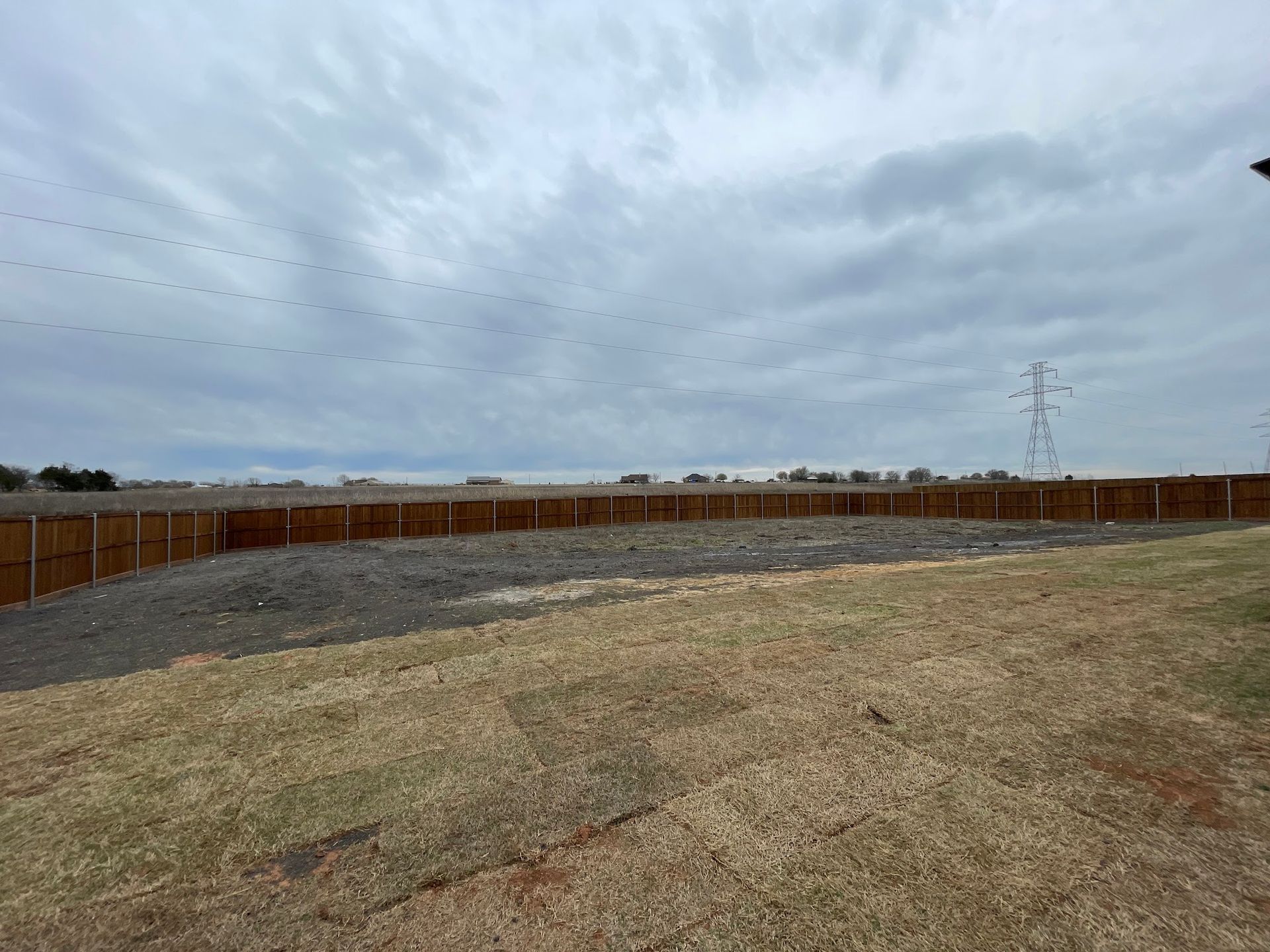 Empty fenced yard under cloudy sky, with gravel and dry grass foreground and power lines in the distance