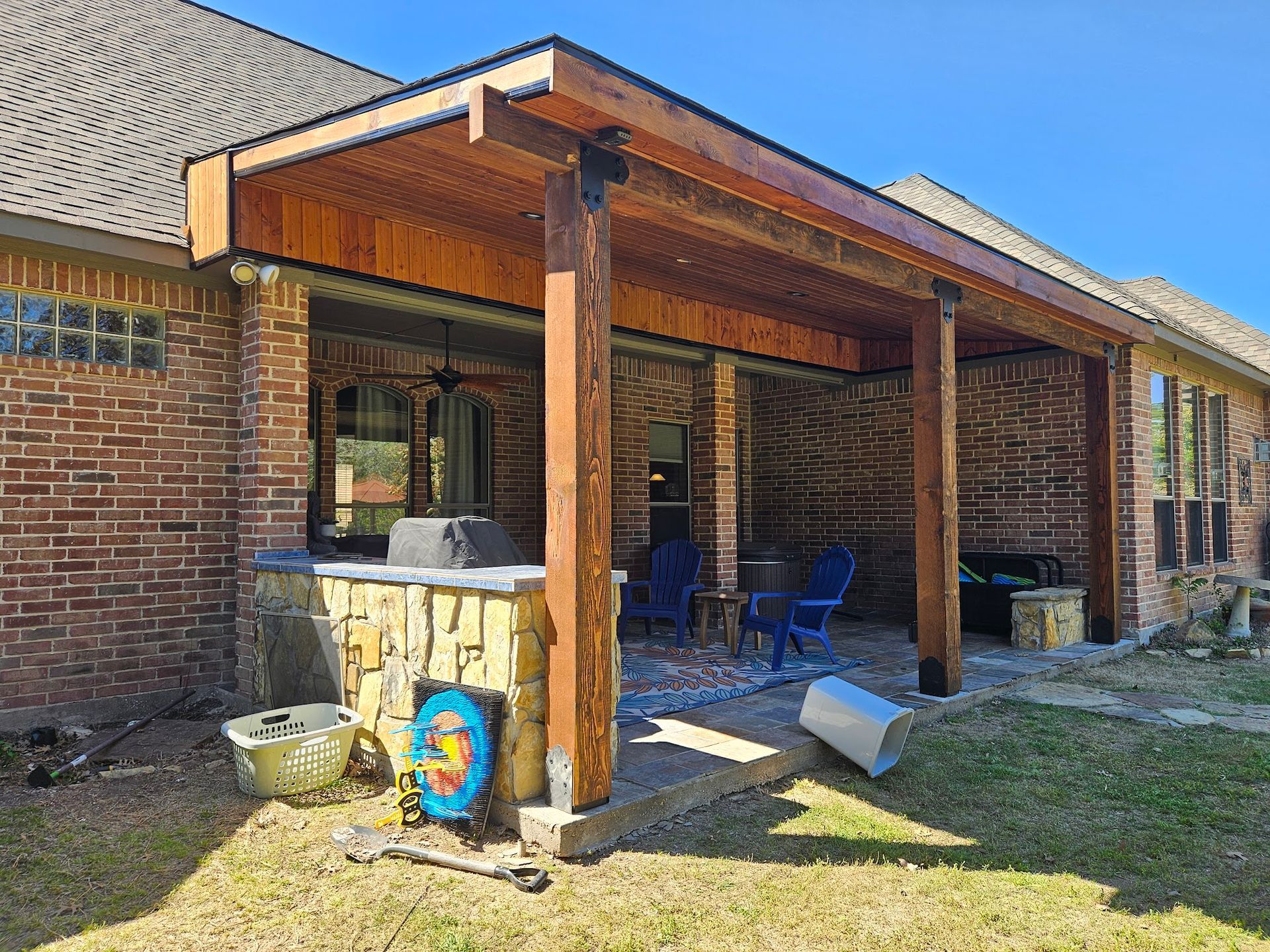 Covered brick patio with wooden posts, blue chairs, and a small stool beside a house