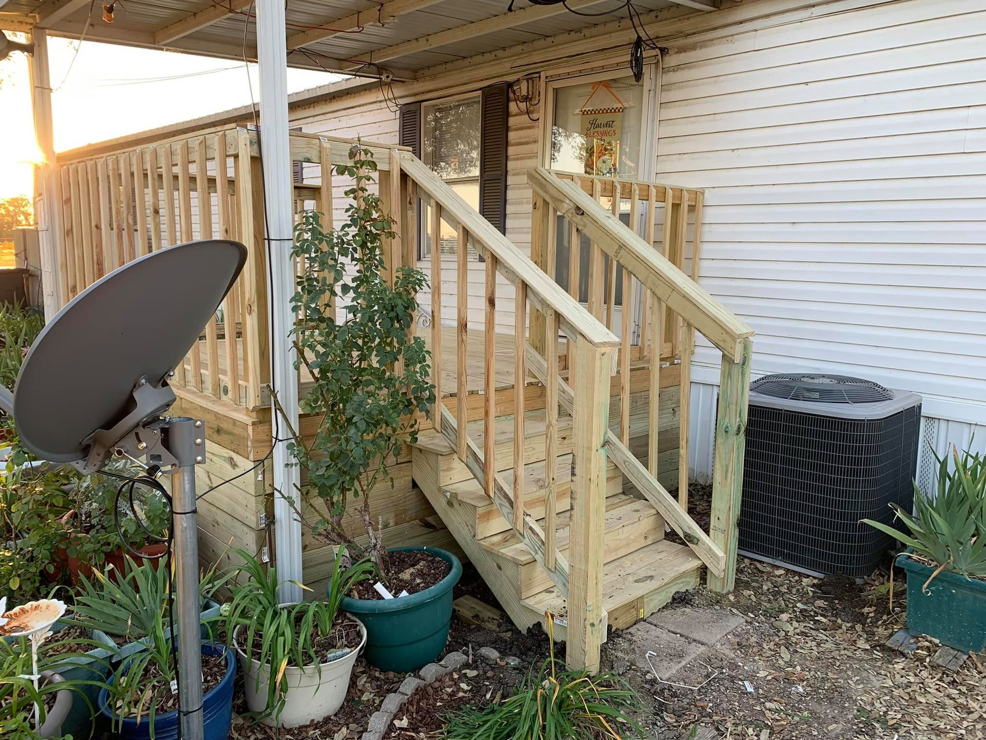 Small wooden porch with stairs, potted plants, a satellite dish, and an air conditioner unit against a white wall.