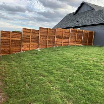 Backyard with a green lawn and a long wooden fence beside a gray house under a cloudy sky