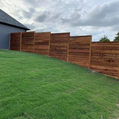 Wooden privacy fence with staggered panels along a sloped green lawn under a cloudy sky