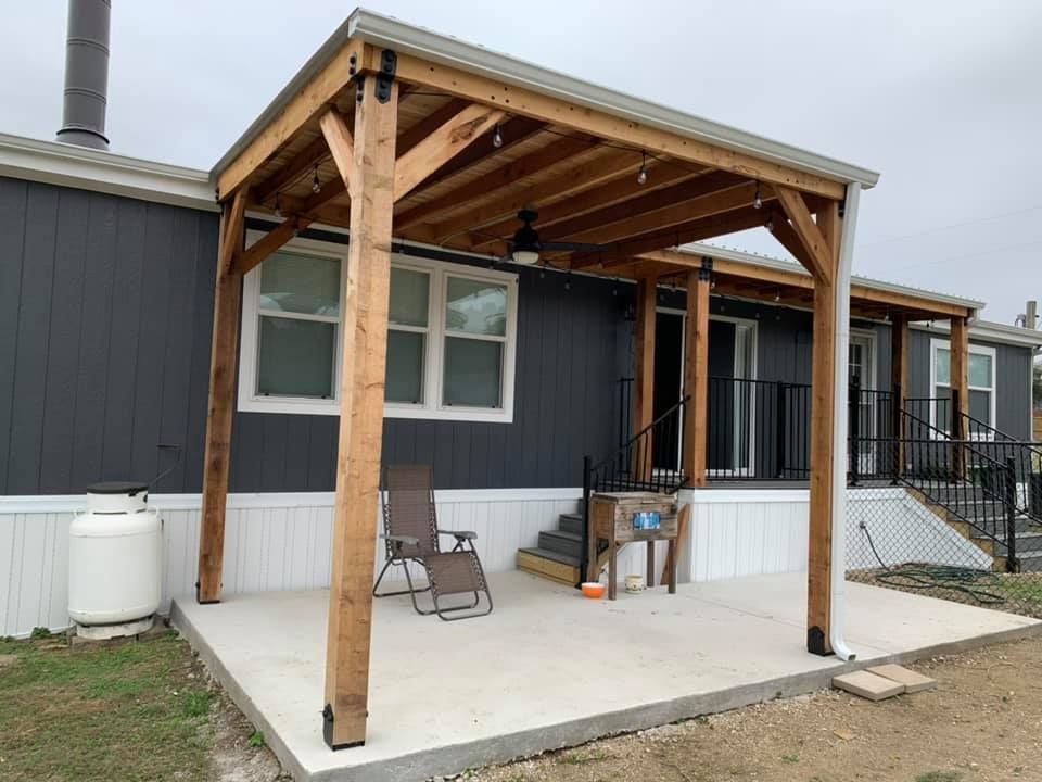 Covered wooden patio with posts and a chair beside a gray mobile home