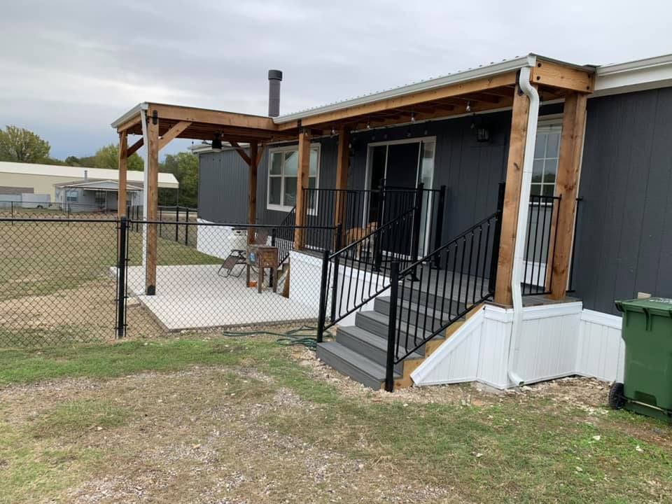 Mobile home porch with black railings, wooden pergola, and steps in a grassy yard