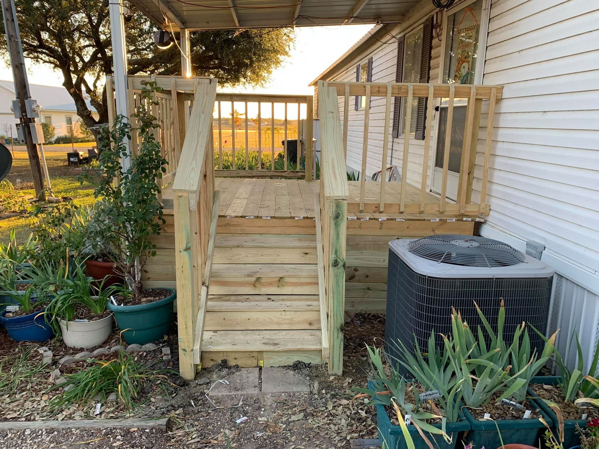 Wooden front steps with railings leading to a porch beside a white house, with potted plants and an HVAC unit nearby.