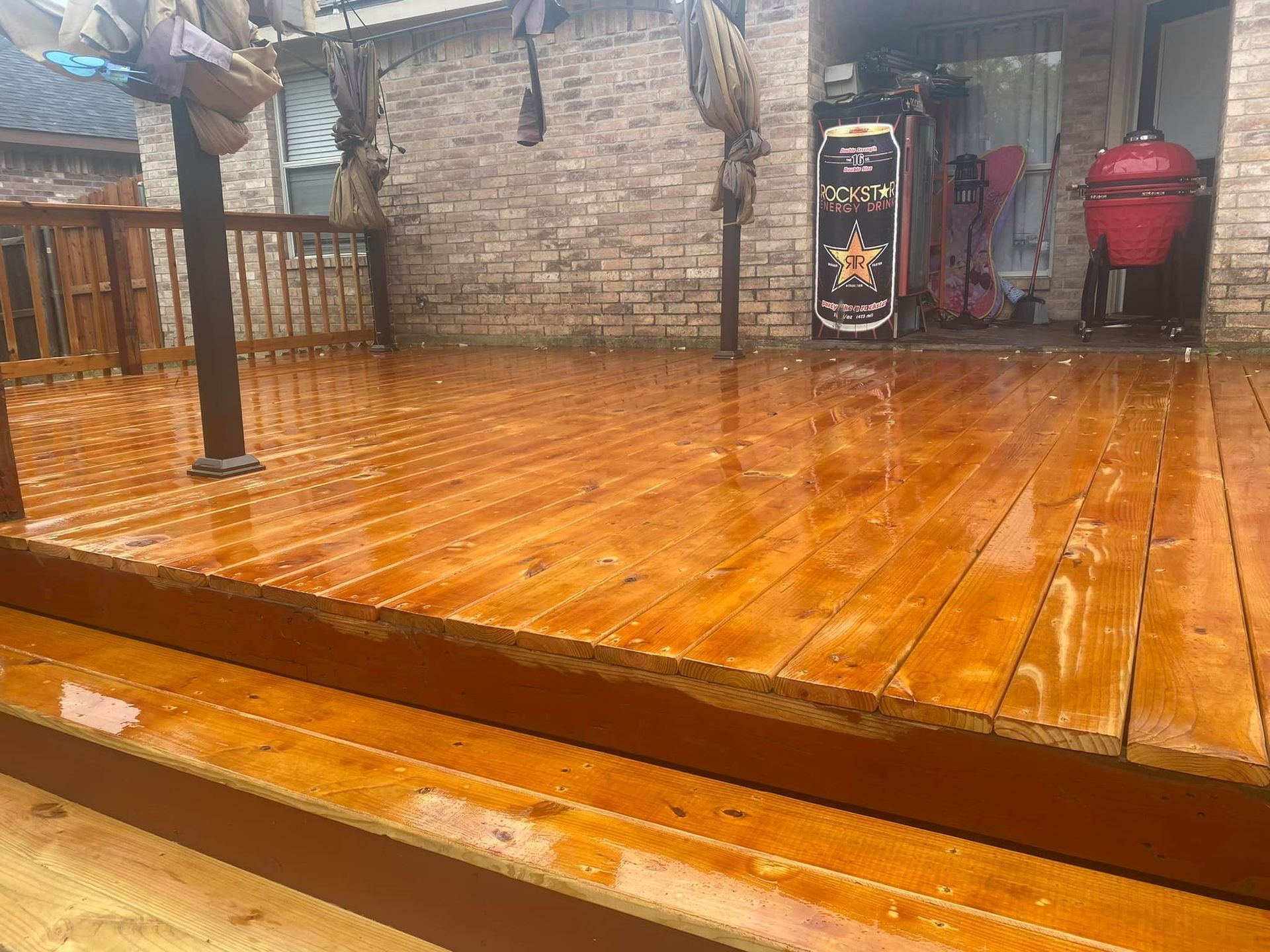 Wet wooden deck beside a brick wall and railing, with a red grill and hanging decor in the background