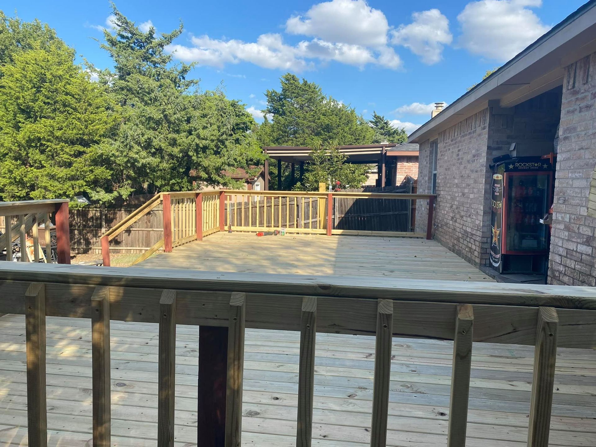 Wooden deck with railing beside a stone building under a blue sky with trees and clouds