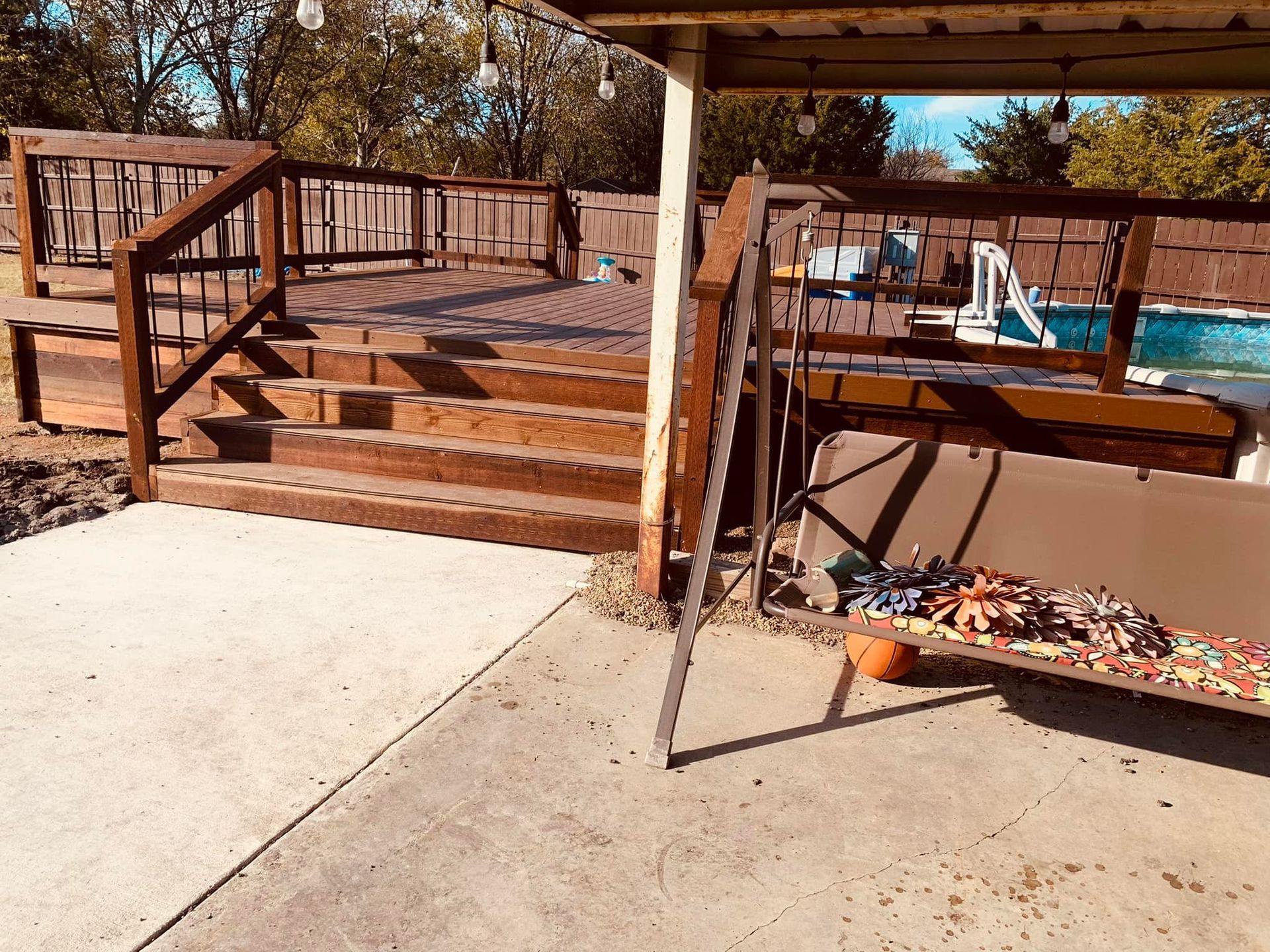 Backyard deck with wooden steps, railing, and a covered pool area beside a concrete patio.