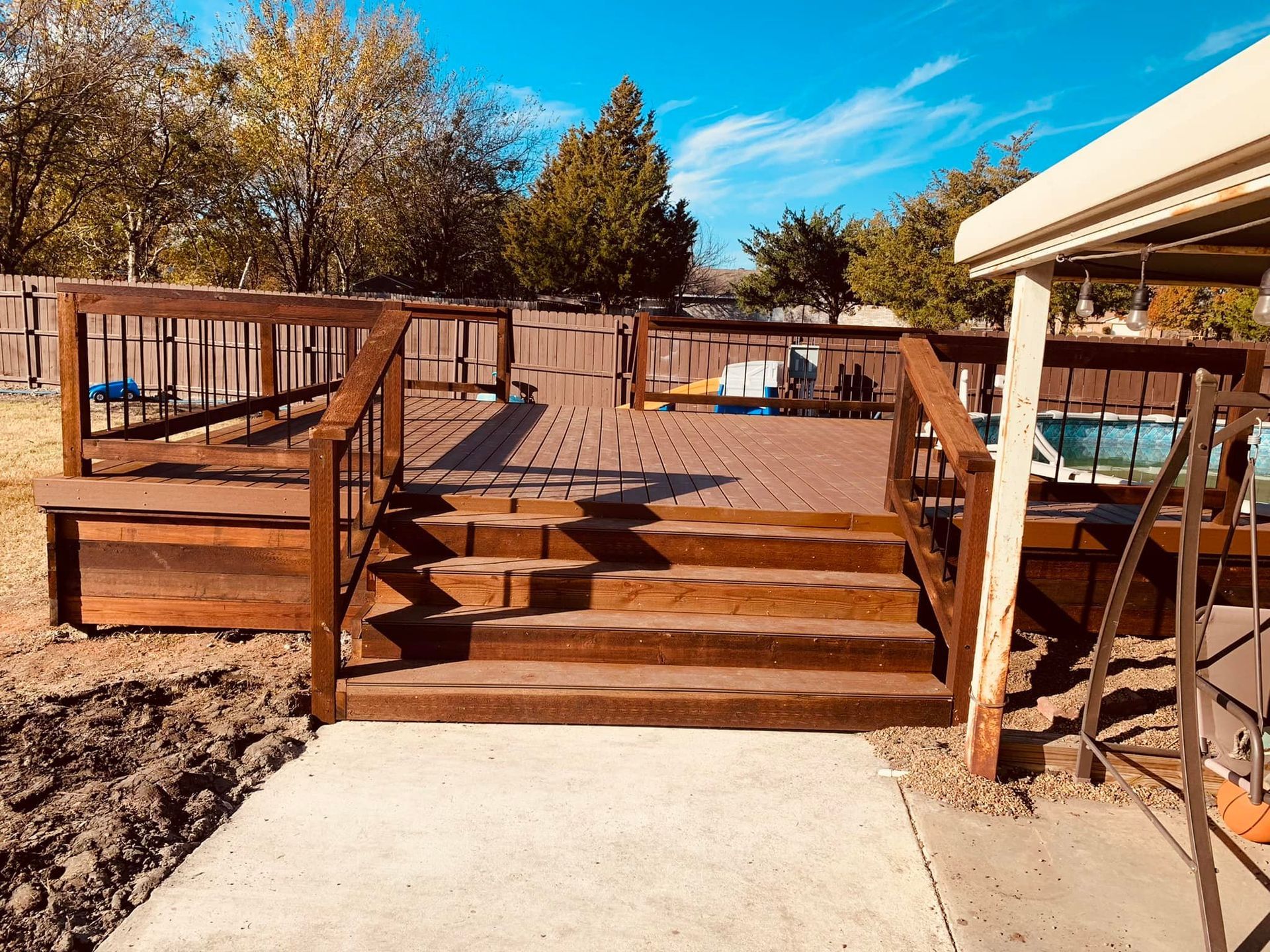 Wooden deck stairs leading to a raised platform in a sunny backyard with fall trees.