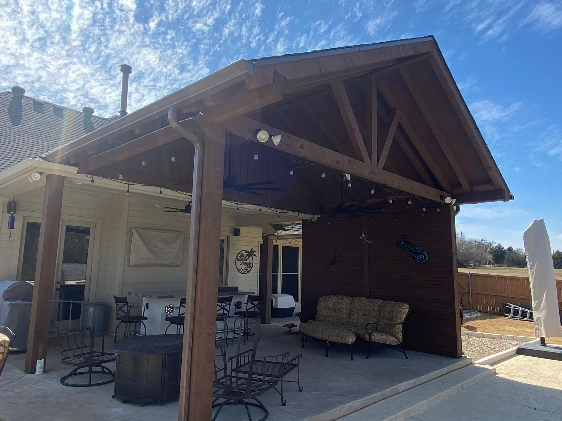 Covered backyard patio with wooden beams, string lights, and outdoor seating beside a brick wall.