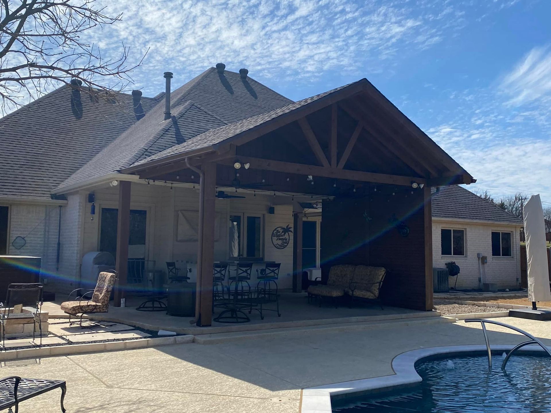Backyard patio with covered seating area beside a swimming pool and a stone house under a blue sky