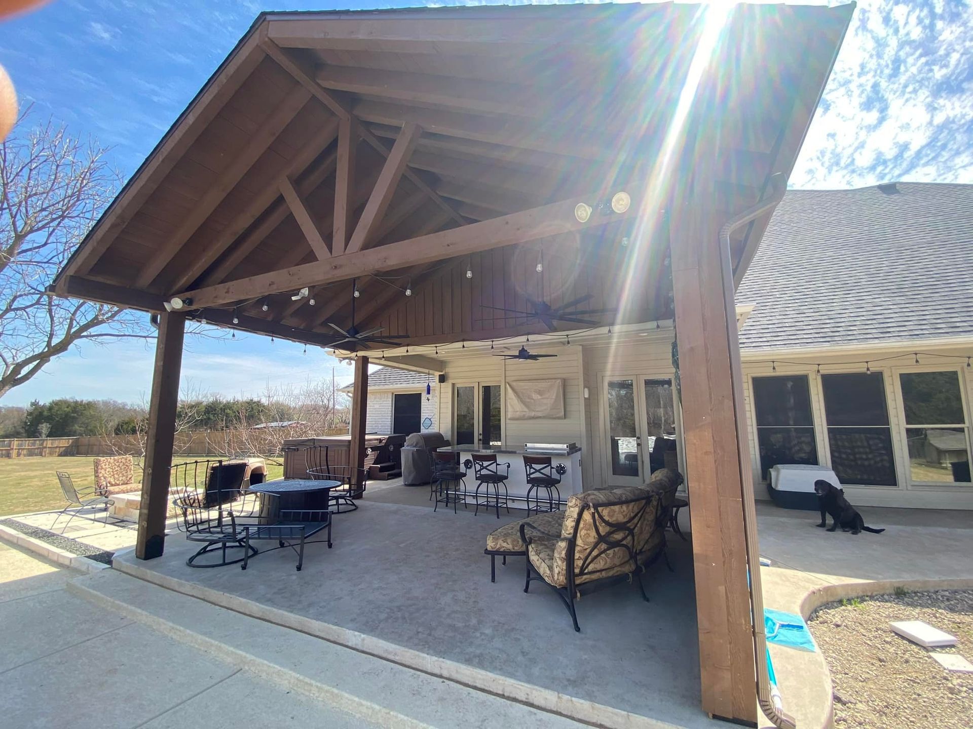 Covered patio with wooden beams, tables and chairs beside a building in bright sunlight.