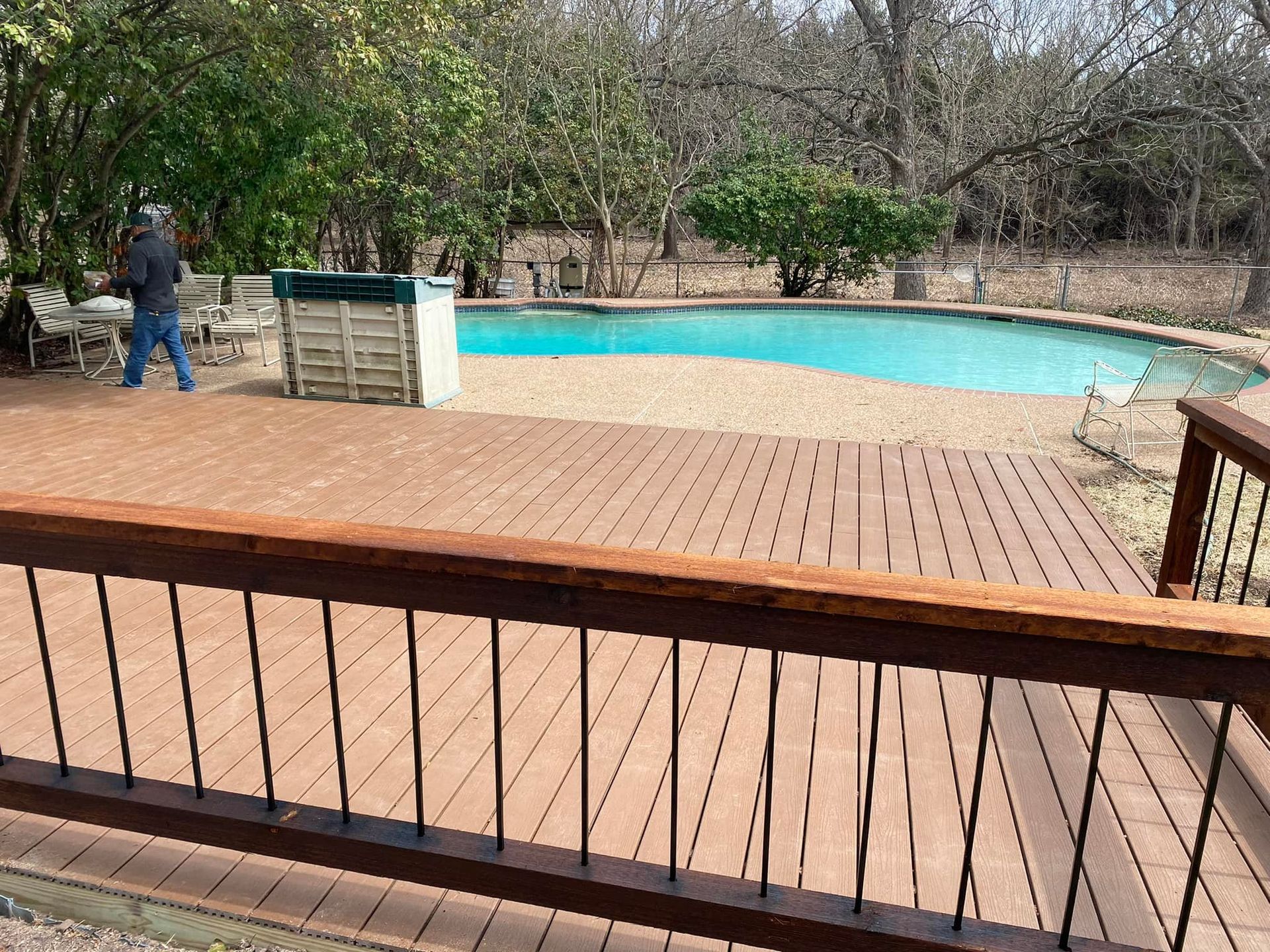 Wooden deck overlooking a turquoise pool with a person walking near the edge in a wooded yard