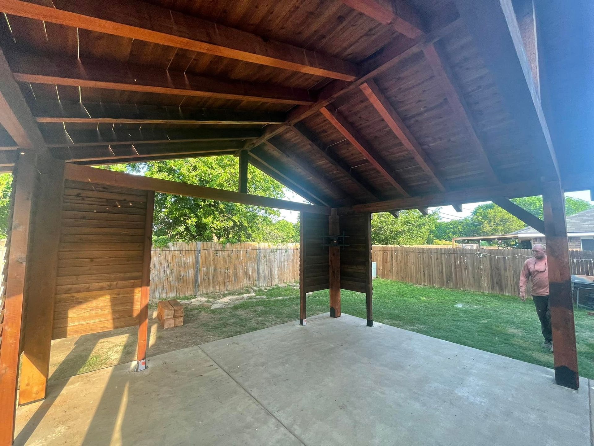 Covered patio with wooden ceiling, concrete floor, and fenced backyard; a person stands at the right edge.