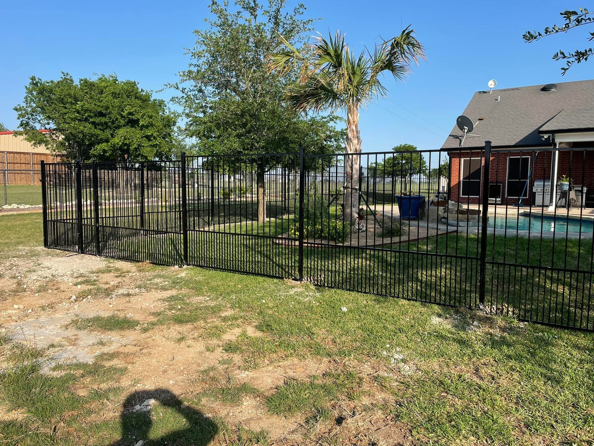 Black metal fence around a backyard with grass, trees, patio, and a house under a blue sky.