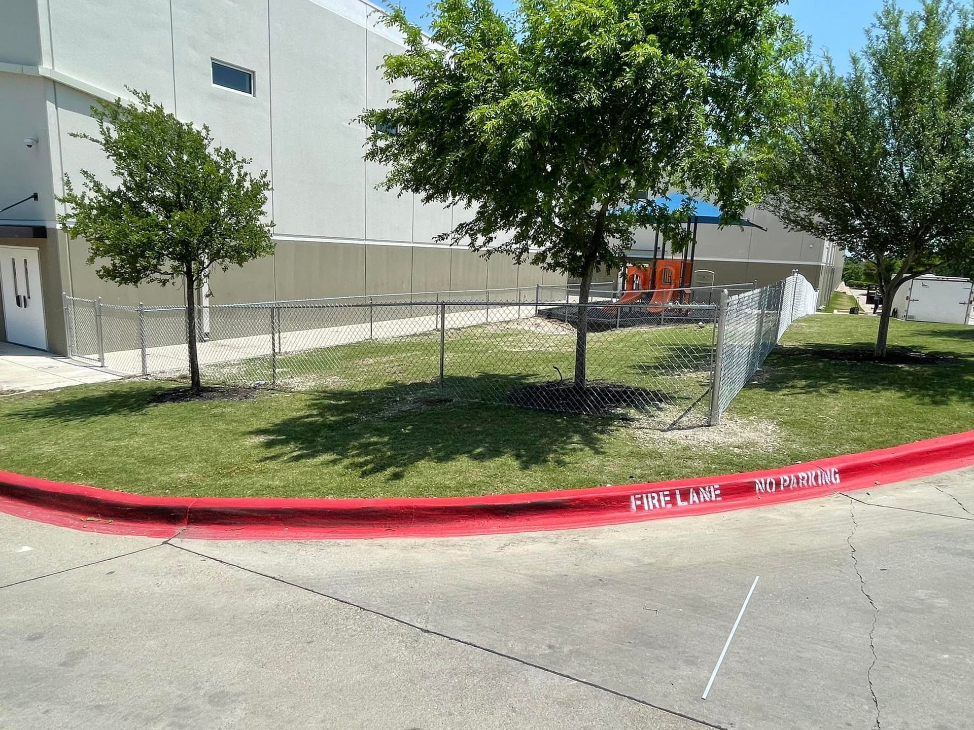 Chain-link fence and trees beside a curb marked “FIRE LANE NO PARKING” outside a building.
