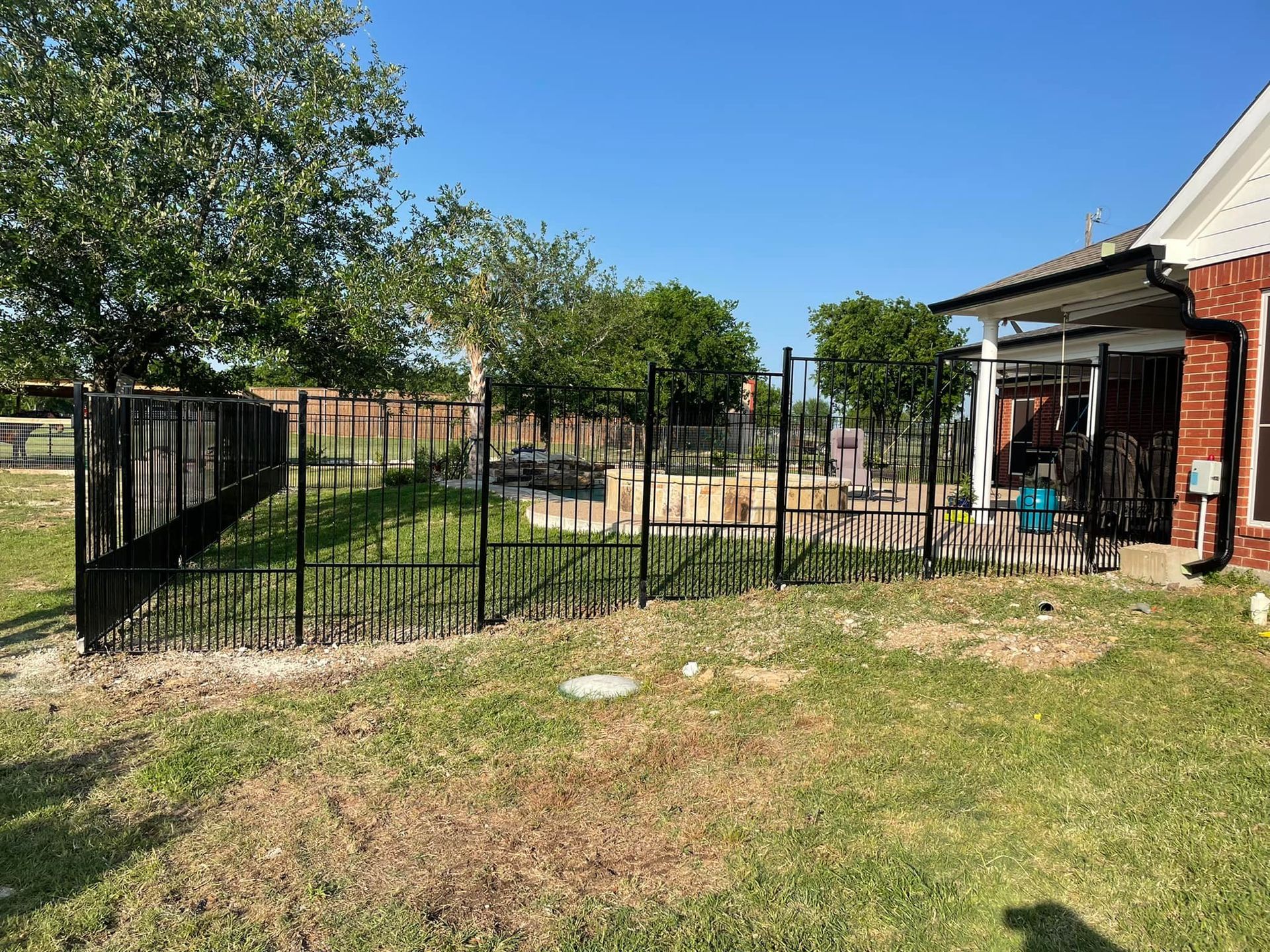 Fenced yard with a grassy lawn, trees, and a brick house under a clear sky