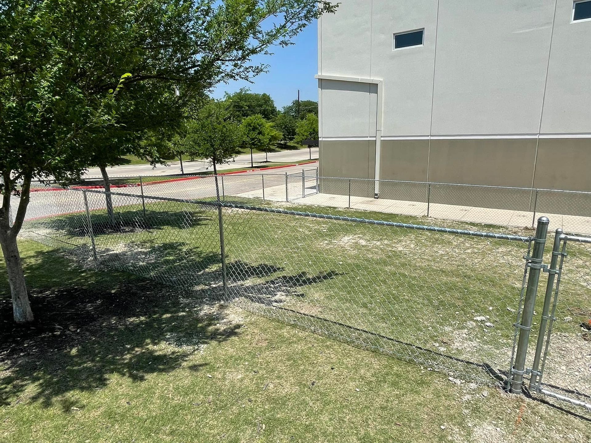 Small fenced grassy yard beside a gray building, with a tree casting shade and a sidewalk beyond.
