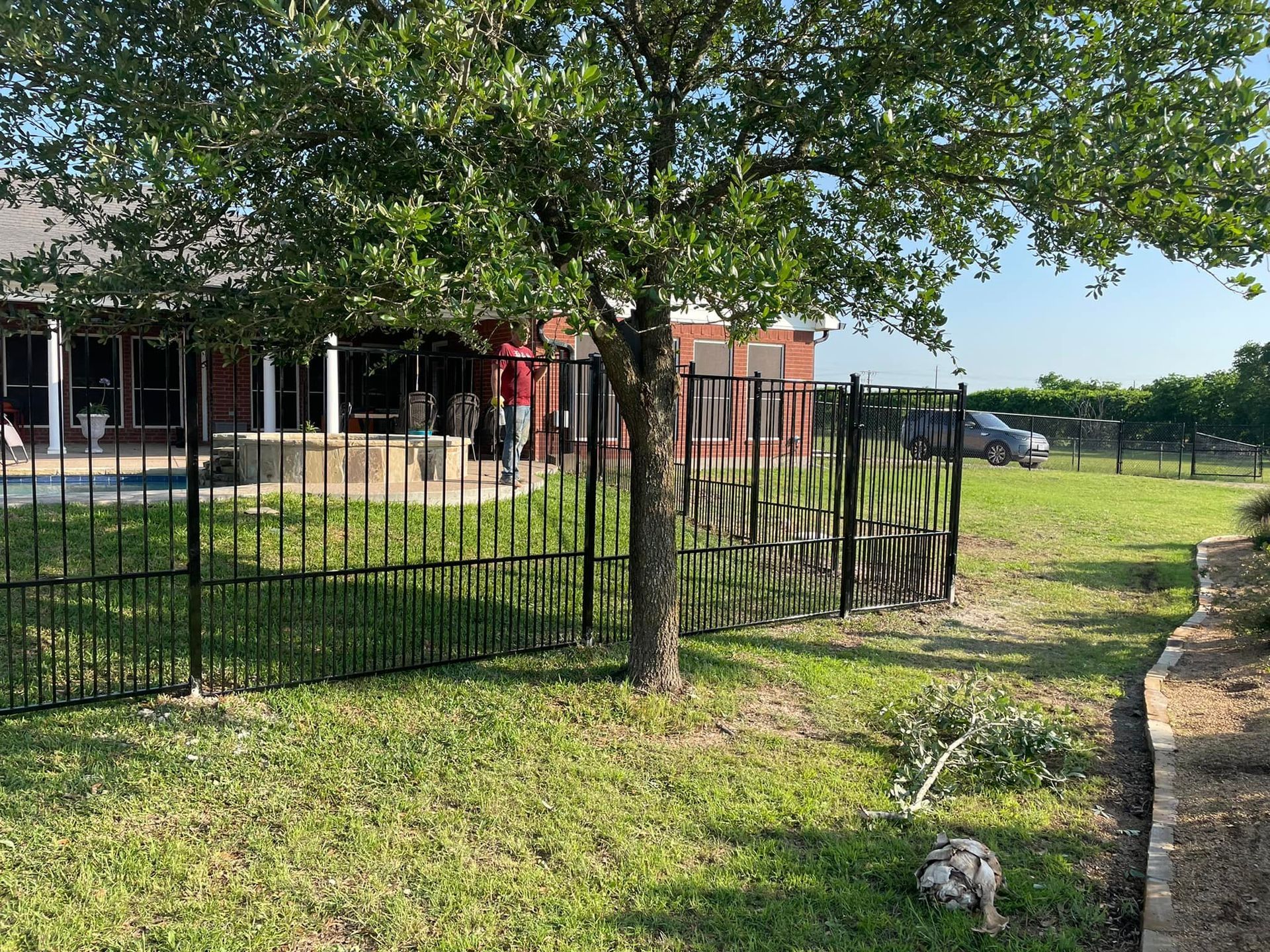 Black metal fence around a grassy yard with a tree, house porch, and parked car in the background.
