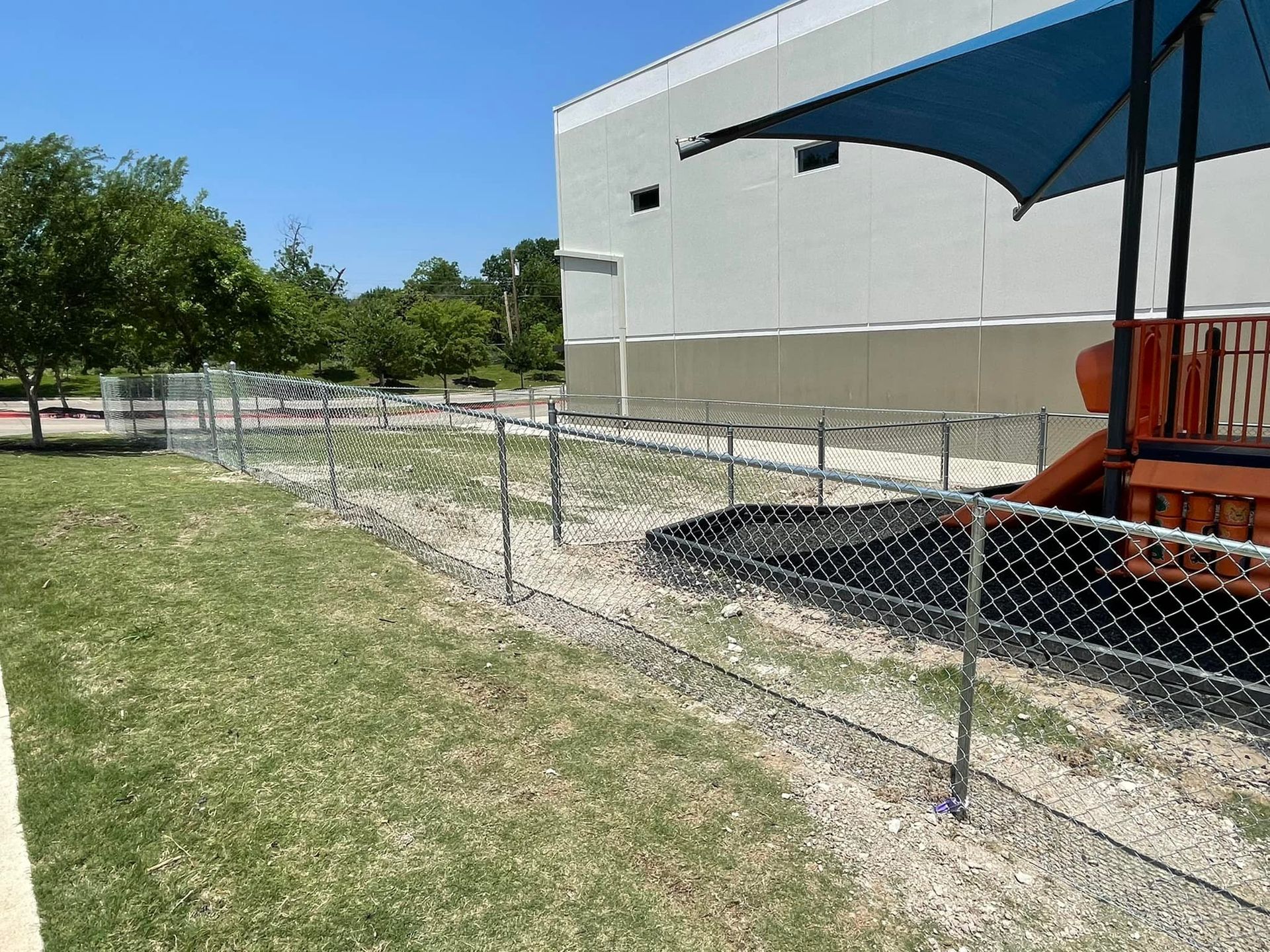 Chain-link fence along a grassy path beside a white building with a blue awning and orange equipment