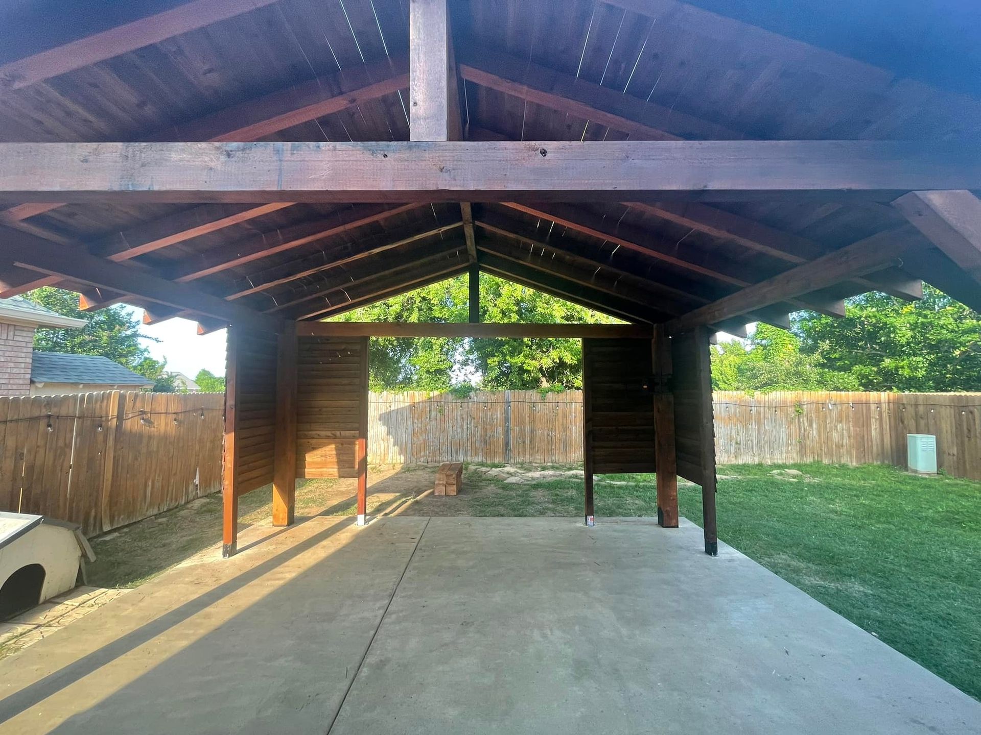 Covered backyard patio with concrete floor, wooden beams, and fenced grass yard.