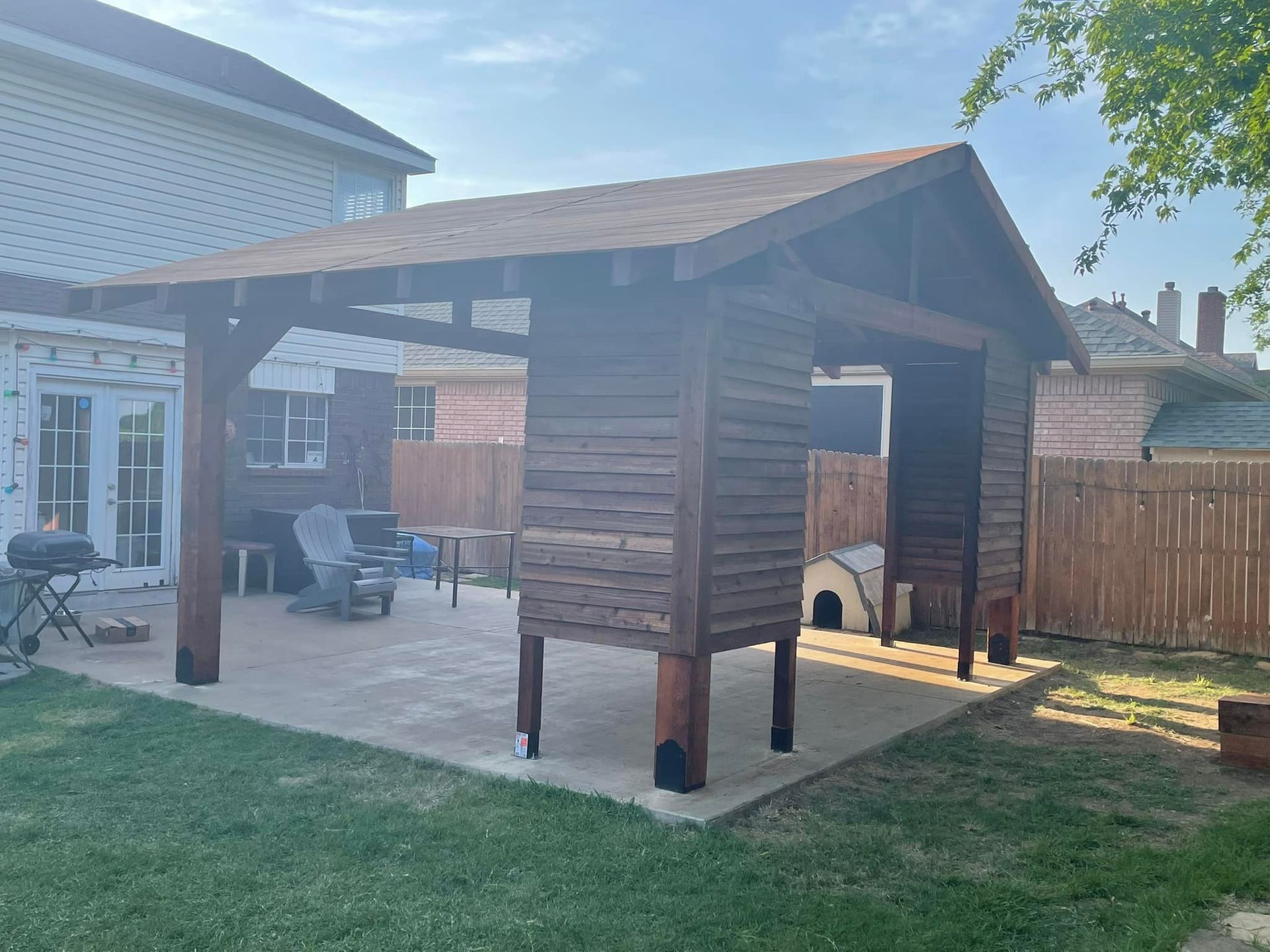Wooden backyard playhouse on stilts beside a house, with a covered porch and fenced yard.