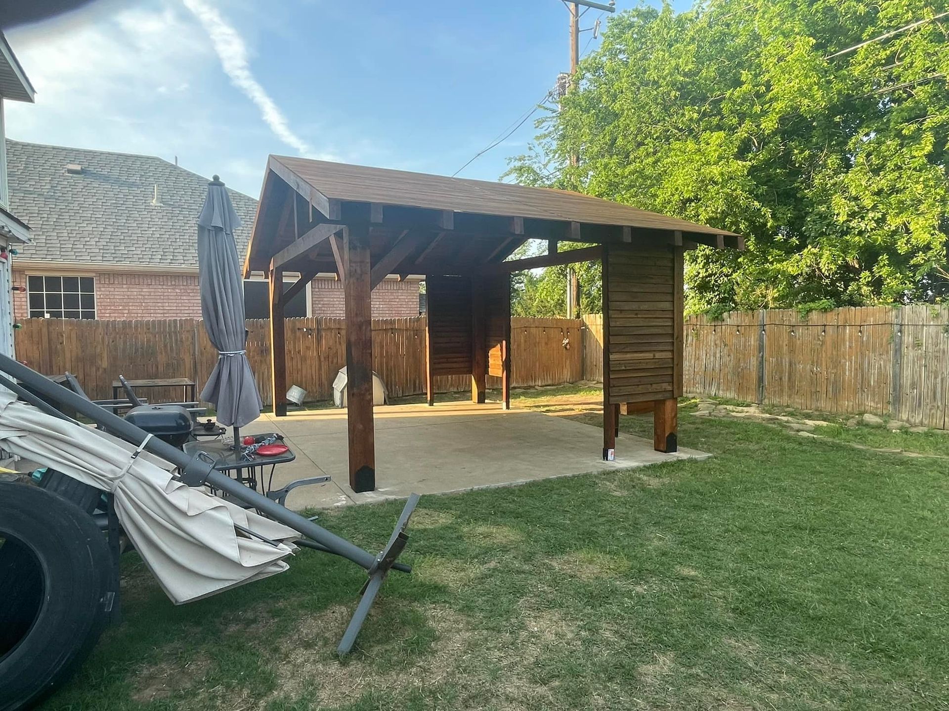 Backyard patio with wooden gazebo, concrete slab, grass, fence, and outdoor furniture under clear sky