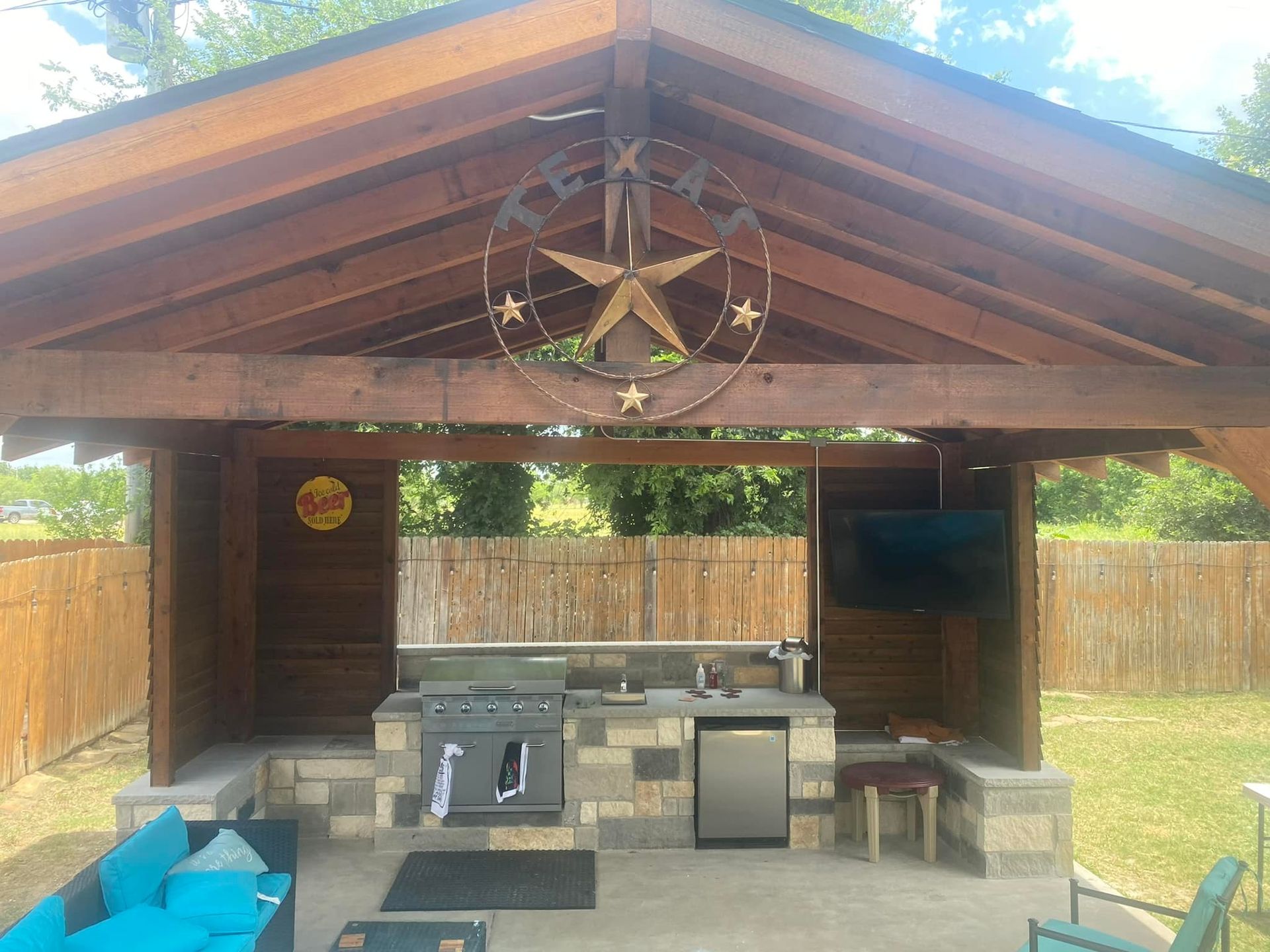 Covered outdoor pavilion with wooden beams, stone fireplace, fridge, chairs, and blue seating under a peaked roof.