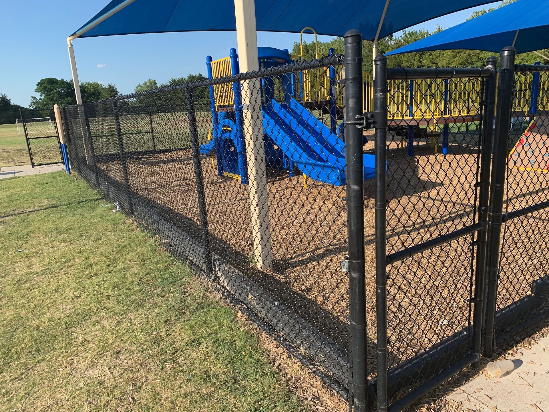 Chain-link fenced playground with blue slides and tan wood chips under a clear sky