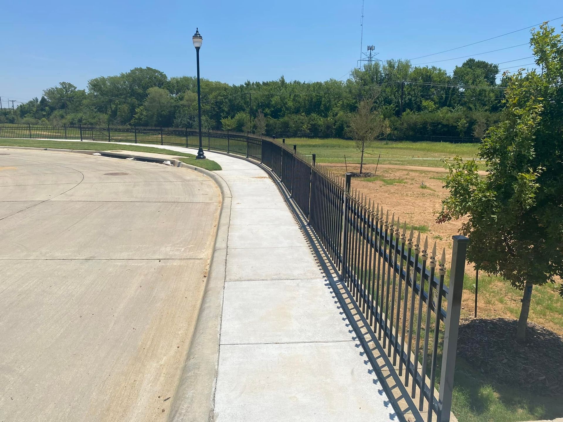 Curved concrete sidewalk beside black metal fence in a sunny park with trees and grass