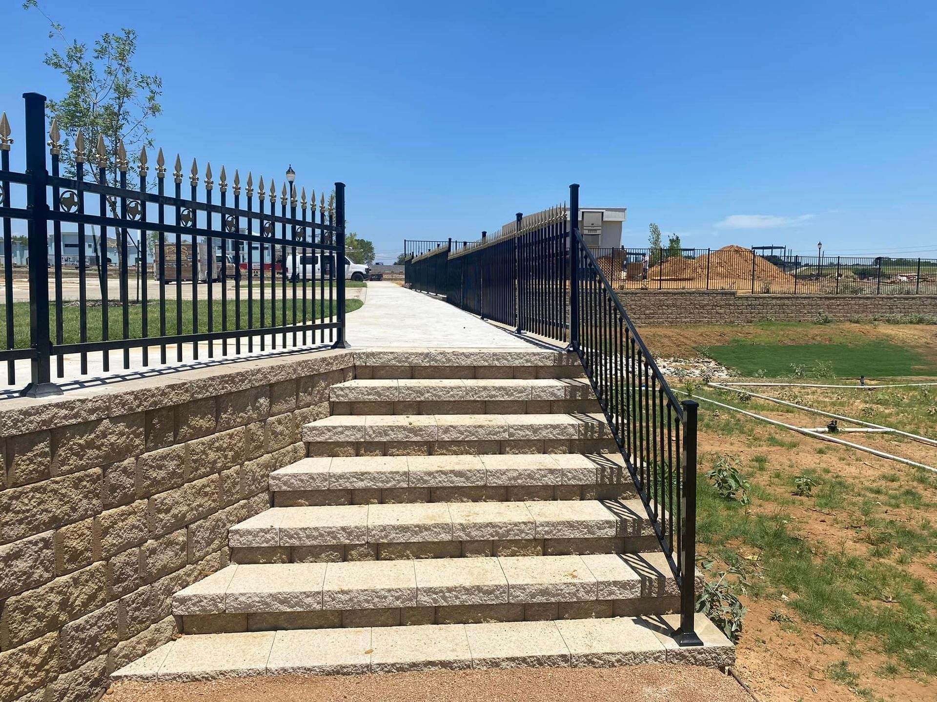 Stone steps with black railings leading up to a paved path beside a grassy park under a blue sky