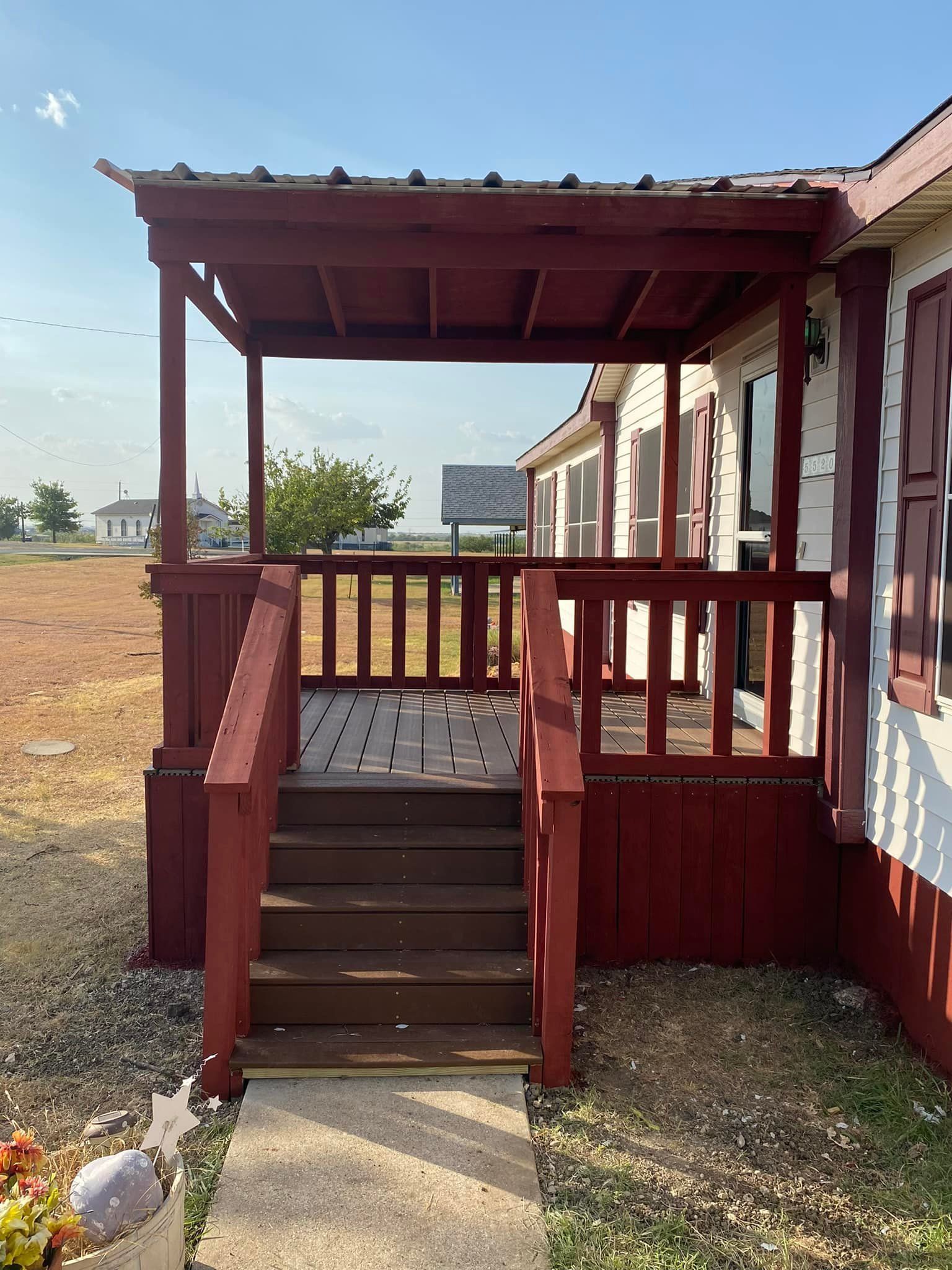 Small red wooden porch with steps and railings under a roof, beside a grassy yard.